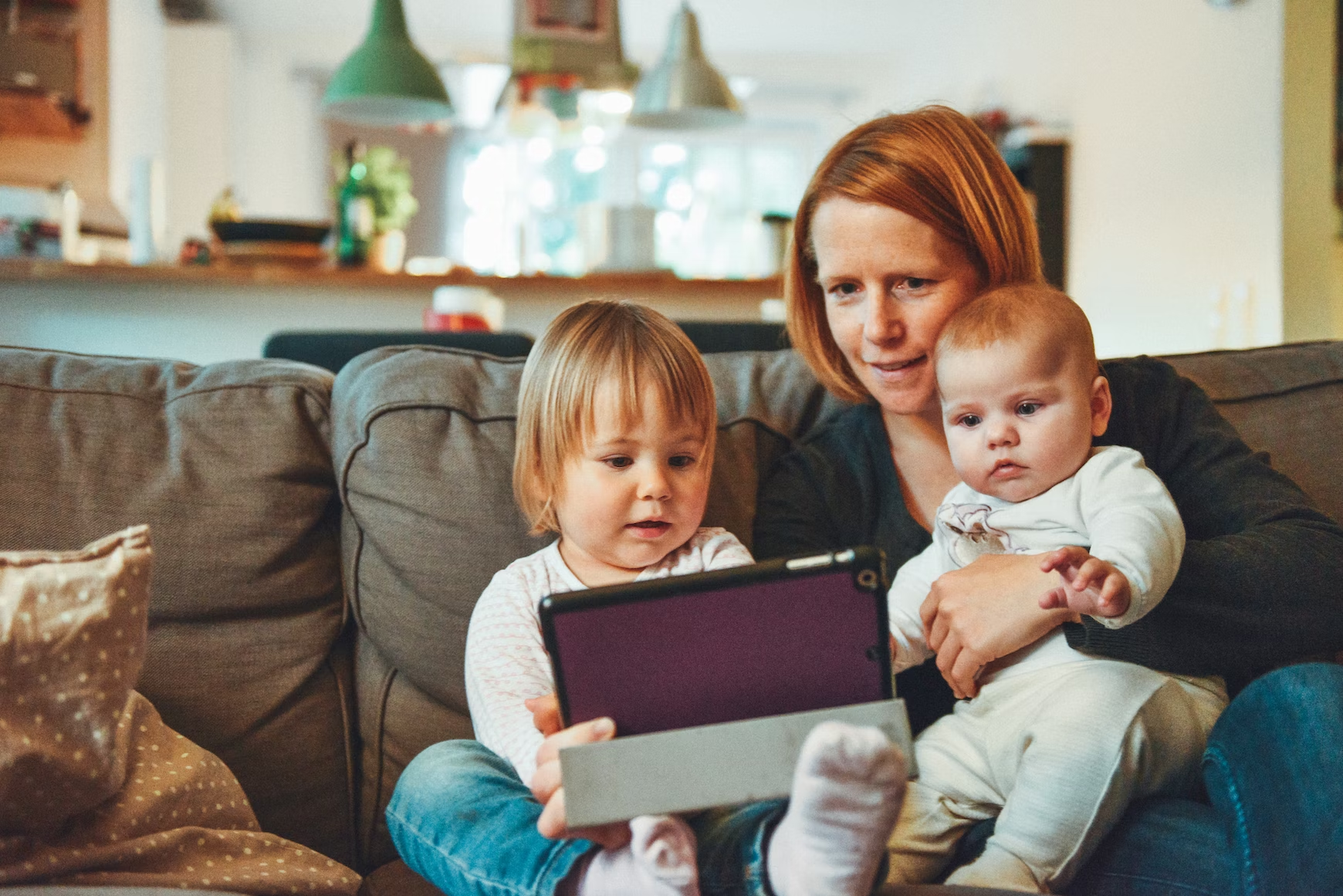 A family with a mom and her two kids sits on the couch looking at a tablet. 